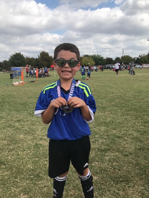 A smiling young soccer player wearing sunglasses holds a medal after playing in an outdoor youth soccer game.